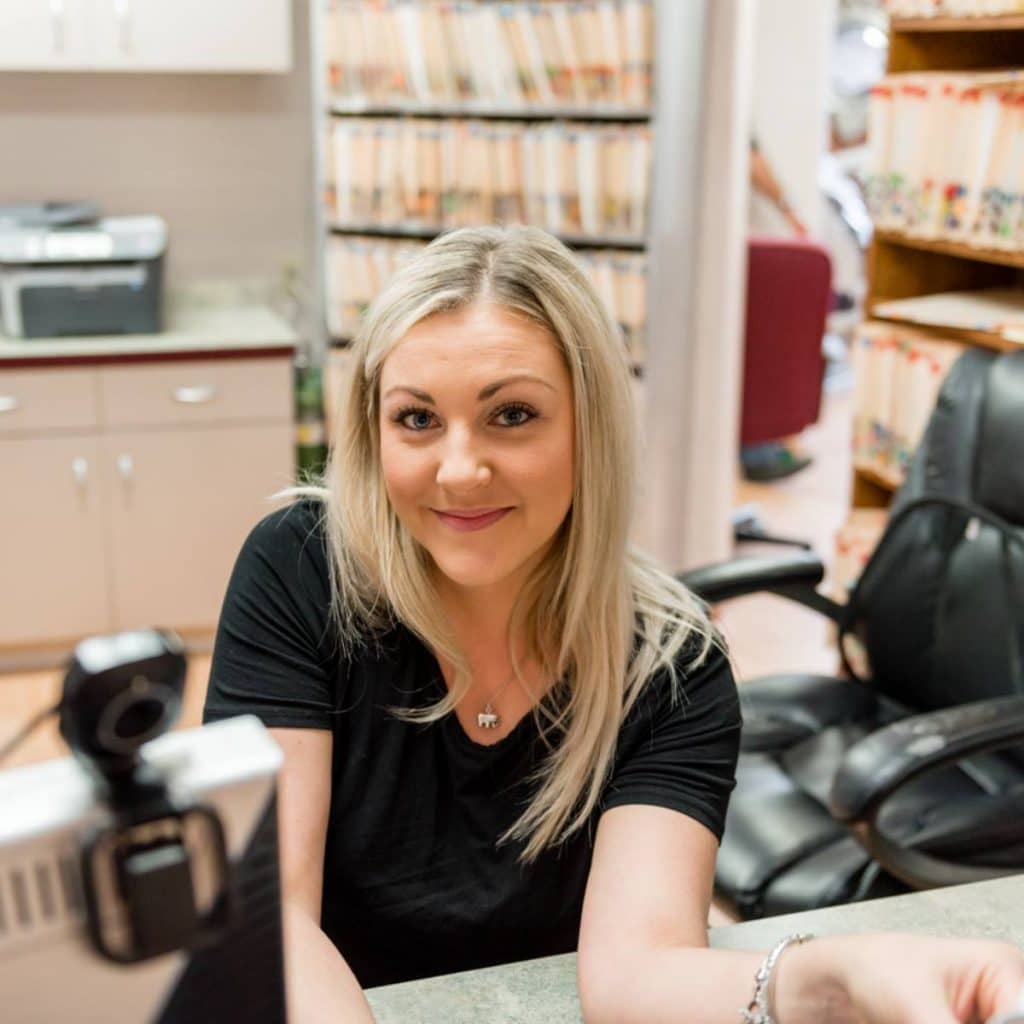 A woman with blonde hair sits at a desk in an office, smiling at the camera. Shelves of files are visible in the background.
