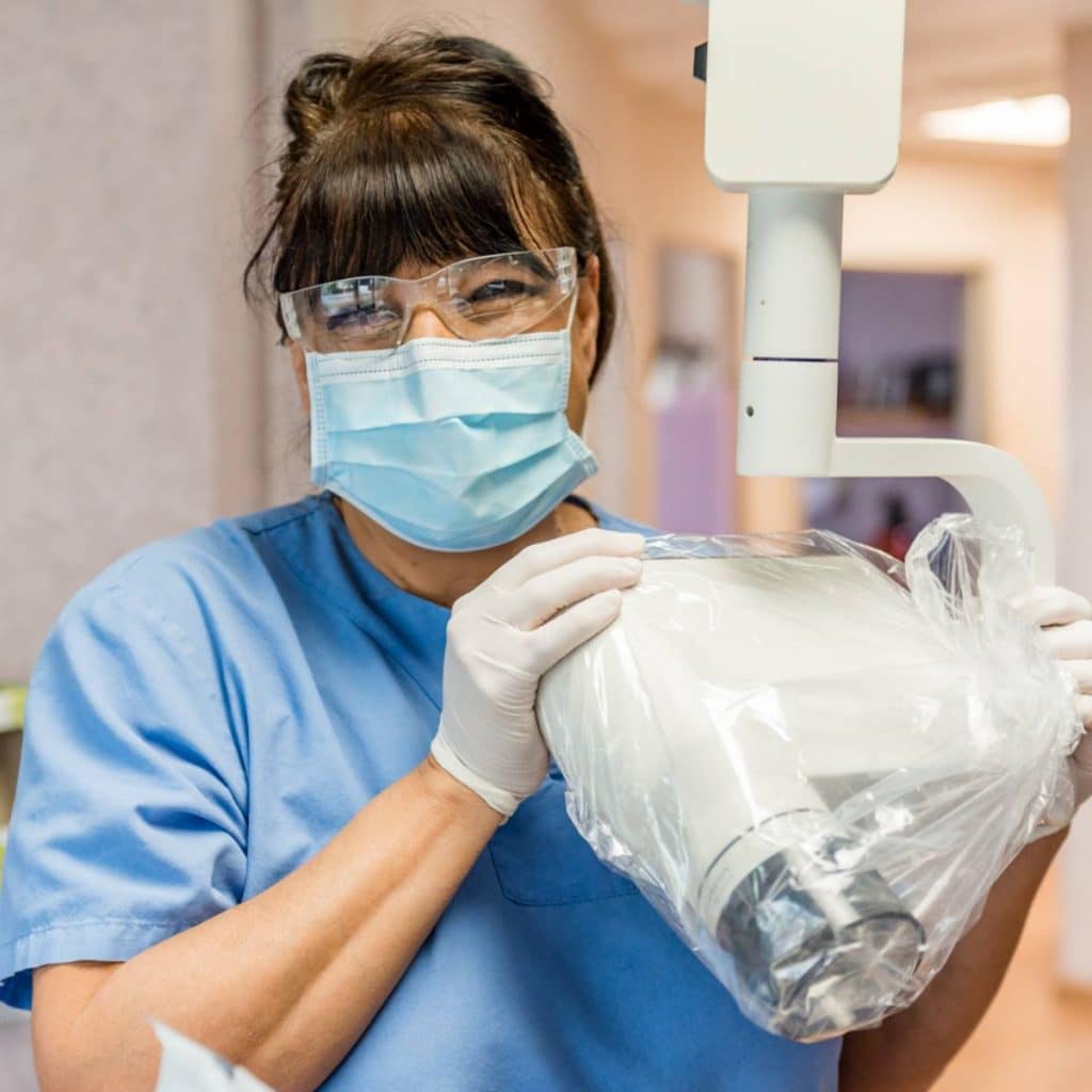 A healthcare worker in scrubs, mask, gloves, and safety glasses holds a covered medical imaging device in a clinical setting.