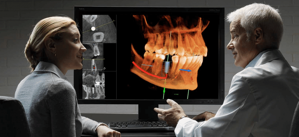 A dentist discusses a 3D dental X-ray with a patient on a computer screen showing jaw and teeth scans.