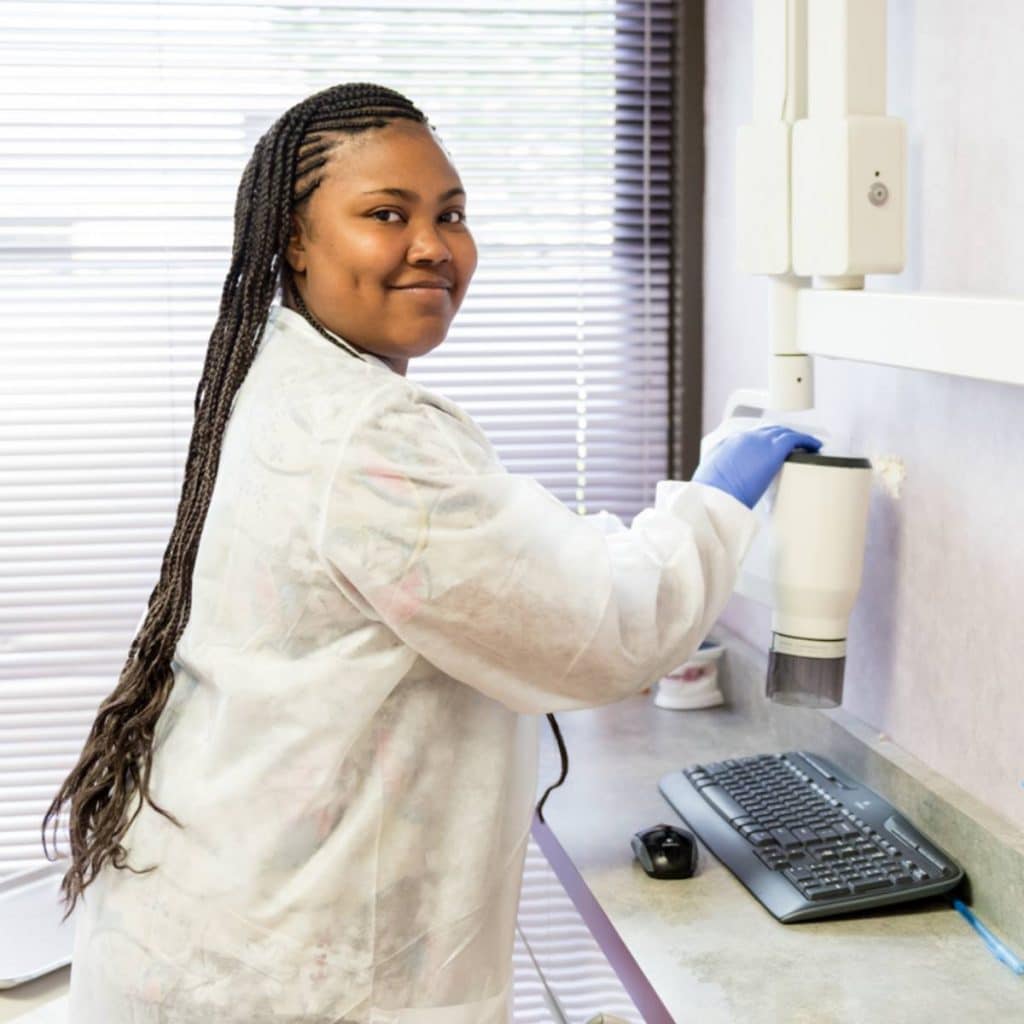 A woman in a lab coat and gloves stands next to dental X-ray equipment, smiling at the camera in a clinical setting.