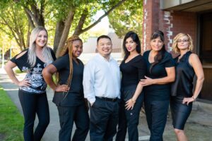 Six adults stand side by side outdoors, five women and one man, posing and smiling at the camera in professional attire. Trees and a brick building are in the background.