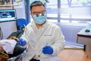 A full service dentist wearing a white coat, mask, and magnifying eyewear holds dental tools in a clinic, with a patient lying on the examination chair.