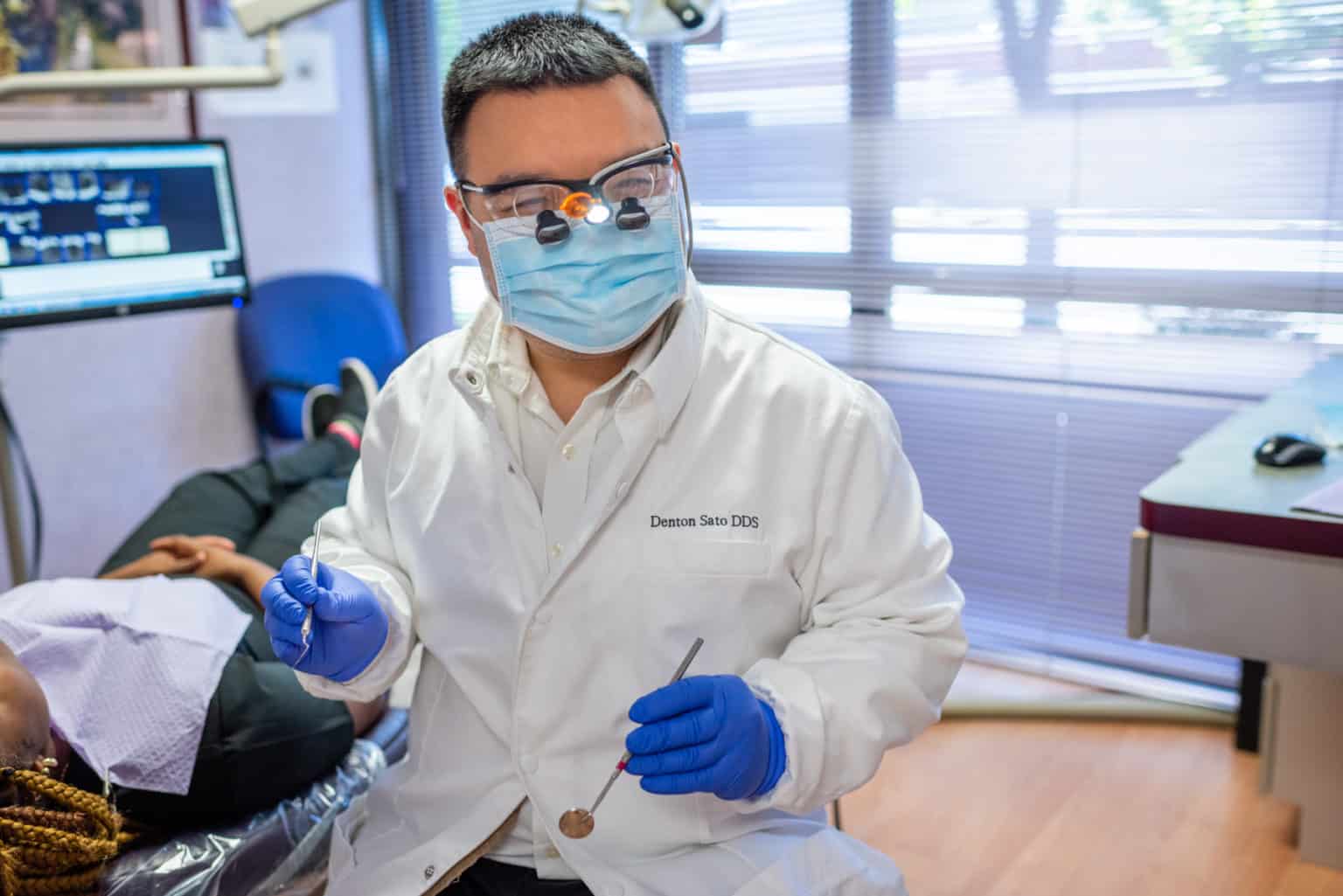 A full service dentist wearing a white coat, mask, and magnifying eyewear holds dental tools in a clinic, with a patient lying on the examination chair.