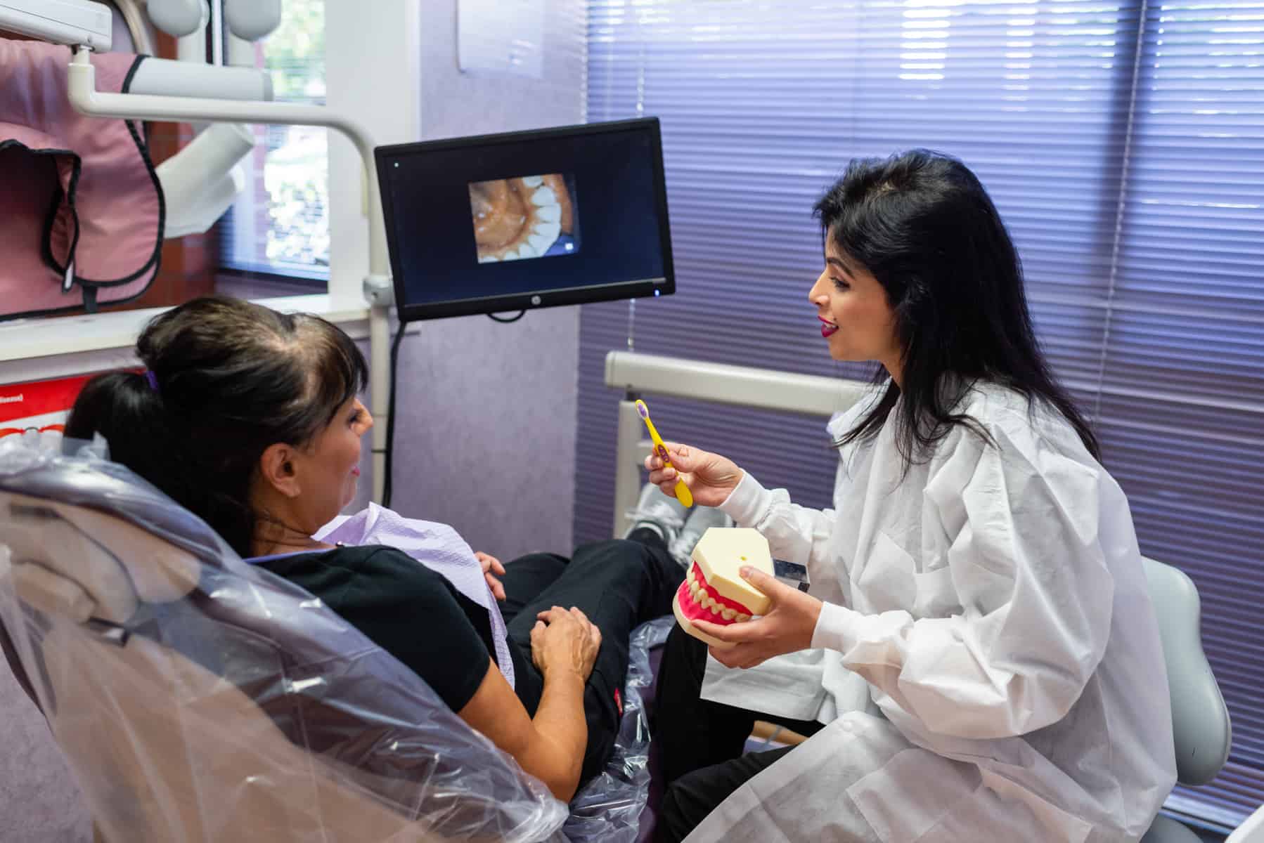 A dentist in a white coat demonstrates tooth brushing on a dental model to a patient sitting in a dental chair, with a dental image displayed on a nearby monitor.