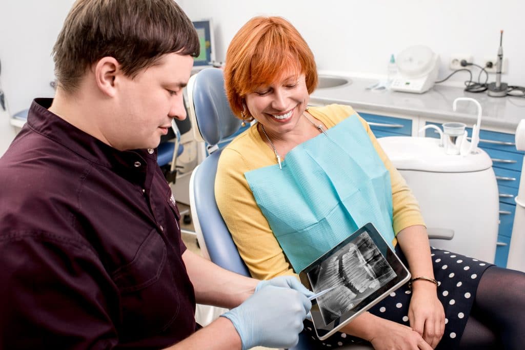 A dentist shows a dental X-ray to a smiling female patient sitting in a dental chair.