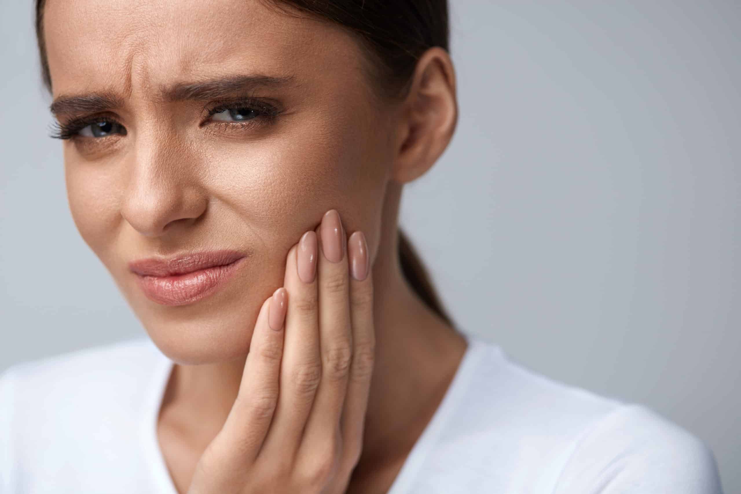 Woman in a white shirt touching her cheek with her hand, appearing to be in pain, possibly due to a toothache—she may need an emergency dentist in Sacramento.