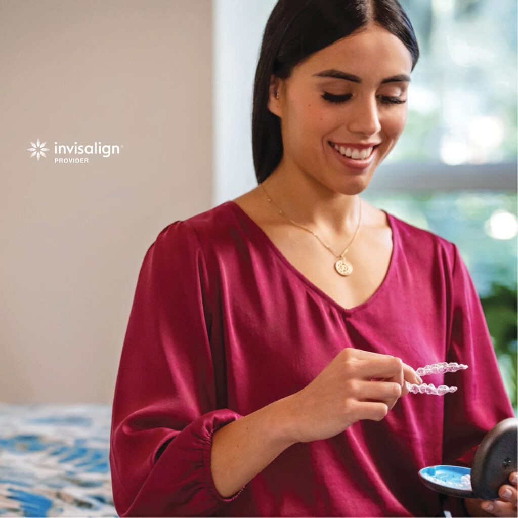 A woman in a maroon blouse holds a clear dental aligner, with an Invisalign case in her other hand. The Invisalign logo appears in the upper left corner.