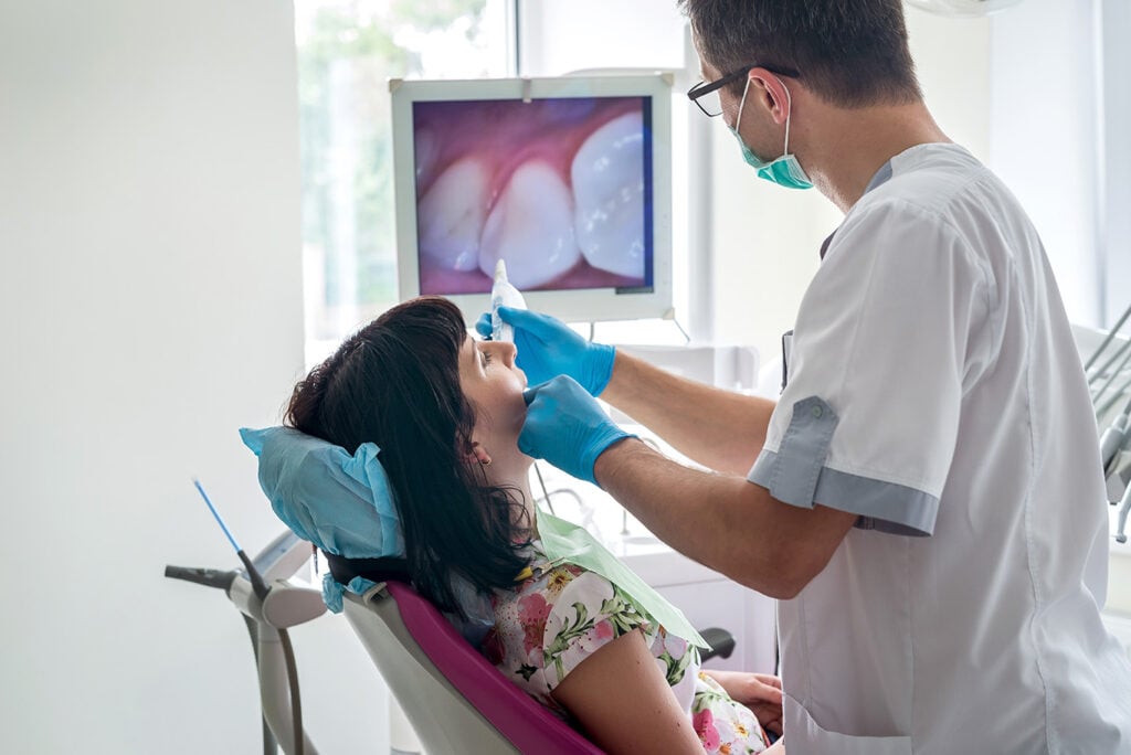 A dentist in gloves and a mask examines a patient's teeth in a dental clinic, with dental diagnostic services displayed on a monitor in the background for precise oral care.