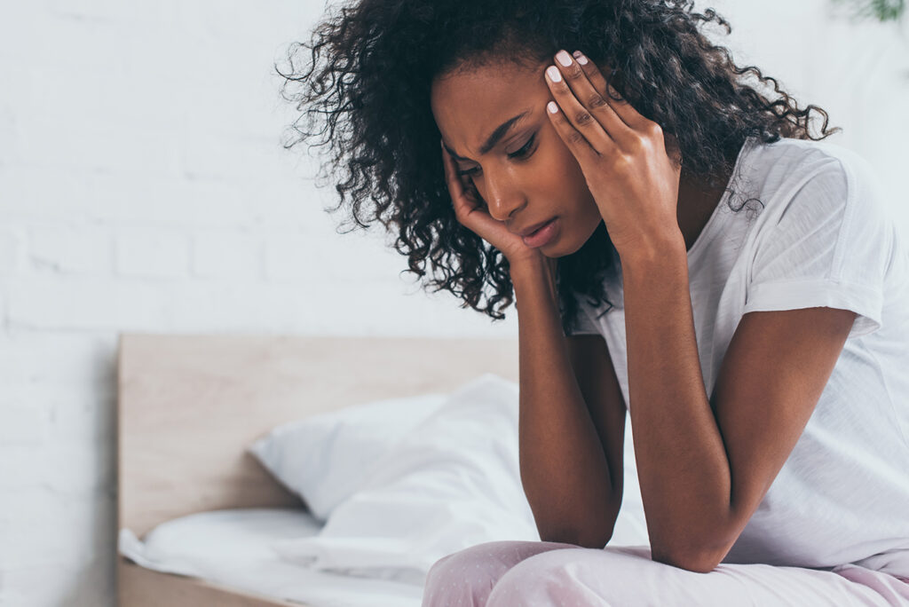 A woman sits on a bed with her hands on her temples, appearing to be in pain or distress, possibly after recent dental medical treatments.