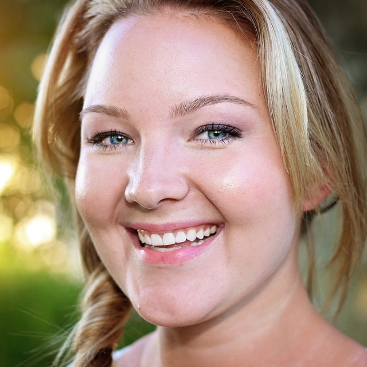 A woman with blonde hair in a side braid smiles at the camera outdoors, her cheerful expression reminiscent of the smile doctor, with a blurred green and yellow background.