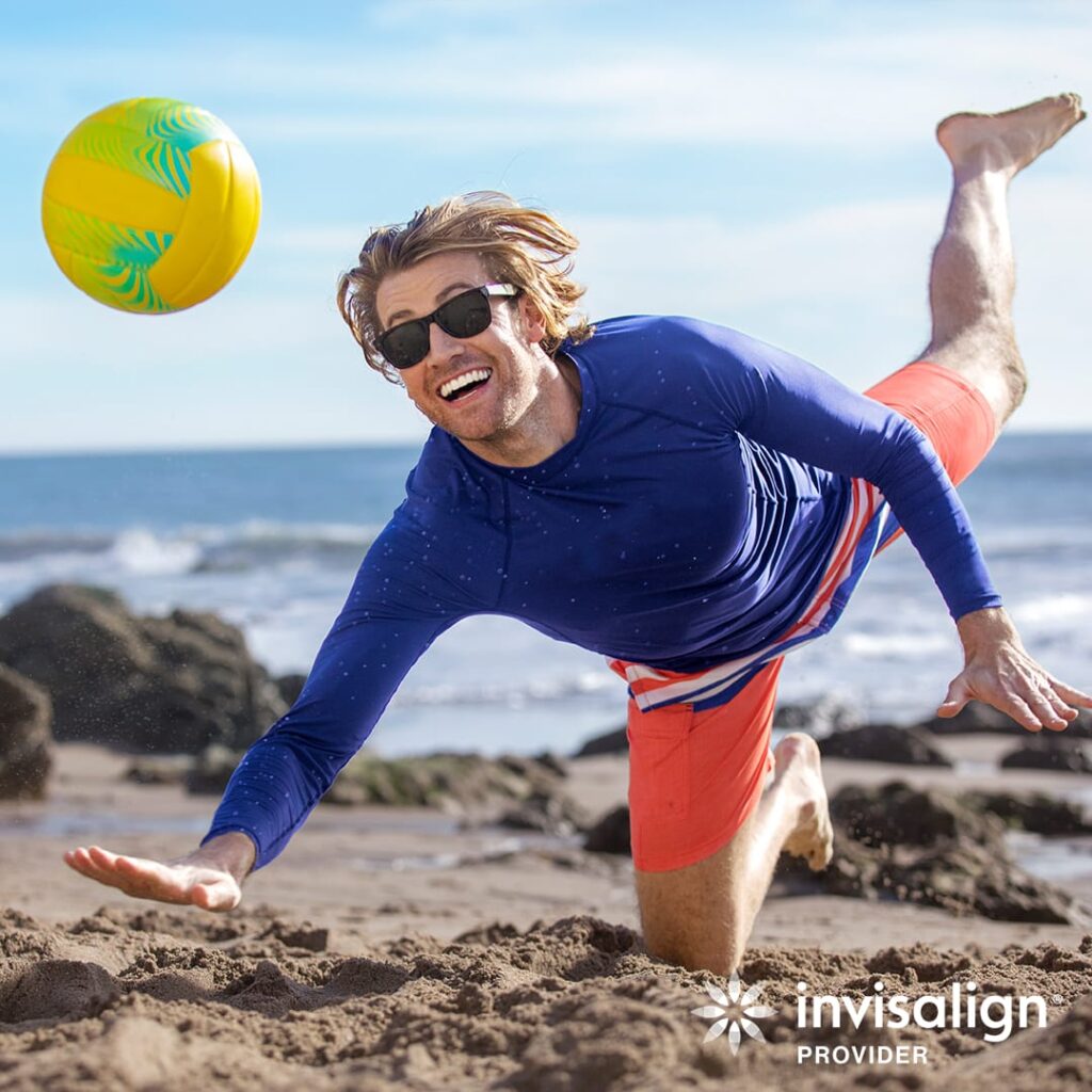 A man in sunglasses and beachwear dives in the sand to hit a volleyball on a sunny beach. The Invisalign Provider logo is in the corner.