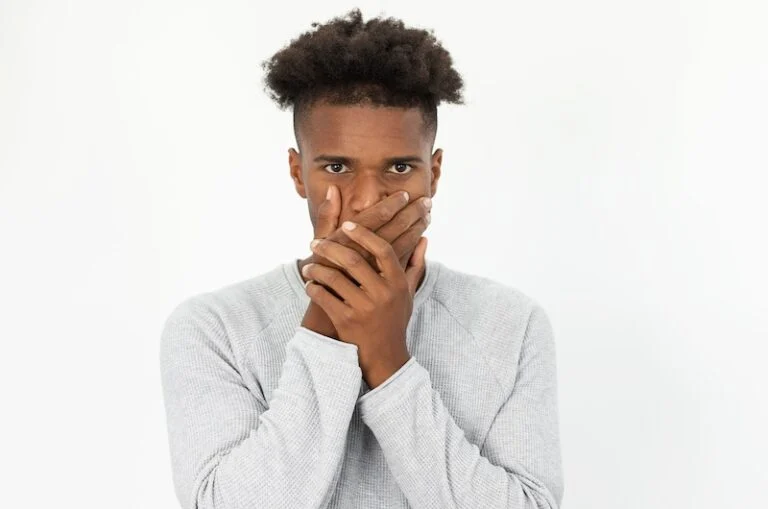 A young man in a gray sweater covers his mouth with both hands, possibly concerned about bacteria or seeking bad breath treatment, while looking directly at the camera against a plain white background.