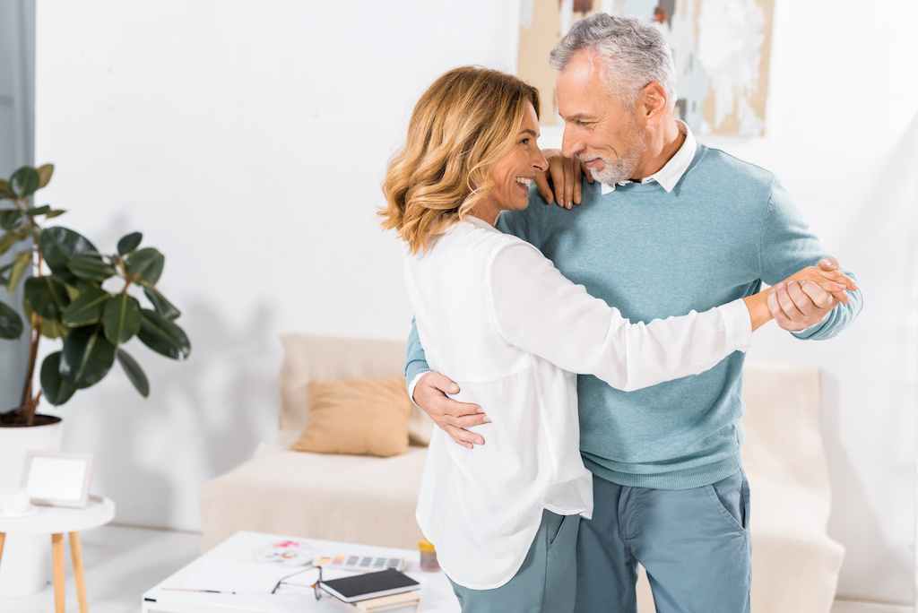 A smiling middle-aged couple dances together in a bright living room, holding each other and making eye contact—embracing the joys of aging and cherishing every moment.