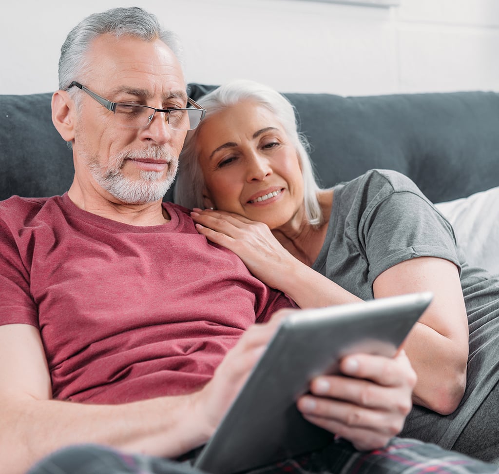 An older couple sits on a sofa together, with the woman resting her head on the man's shoulder while he uses a tablet—his smile showing signs of darkened teeth from age and root decay.