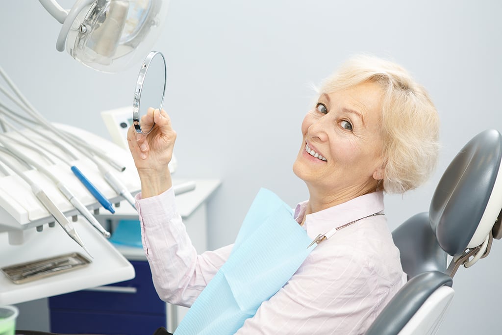 Older woman sitting in a dental chair, holding a small mirror and smiling, with dental equipment visible beside her—highlighting attentive dentistry for seniors.