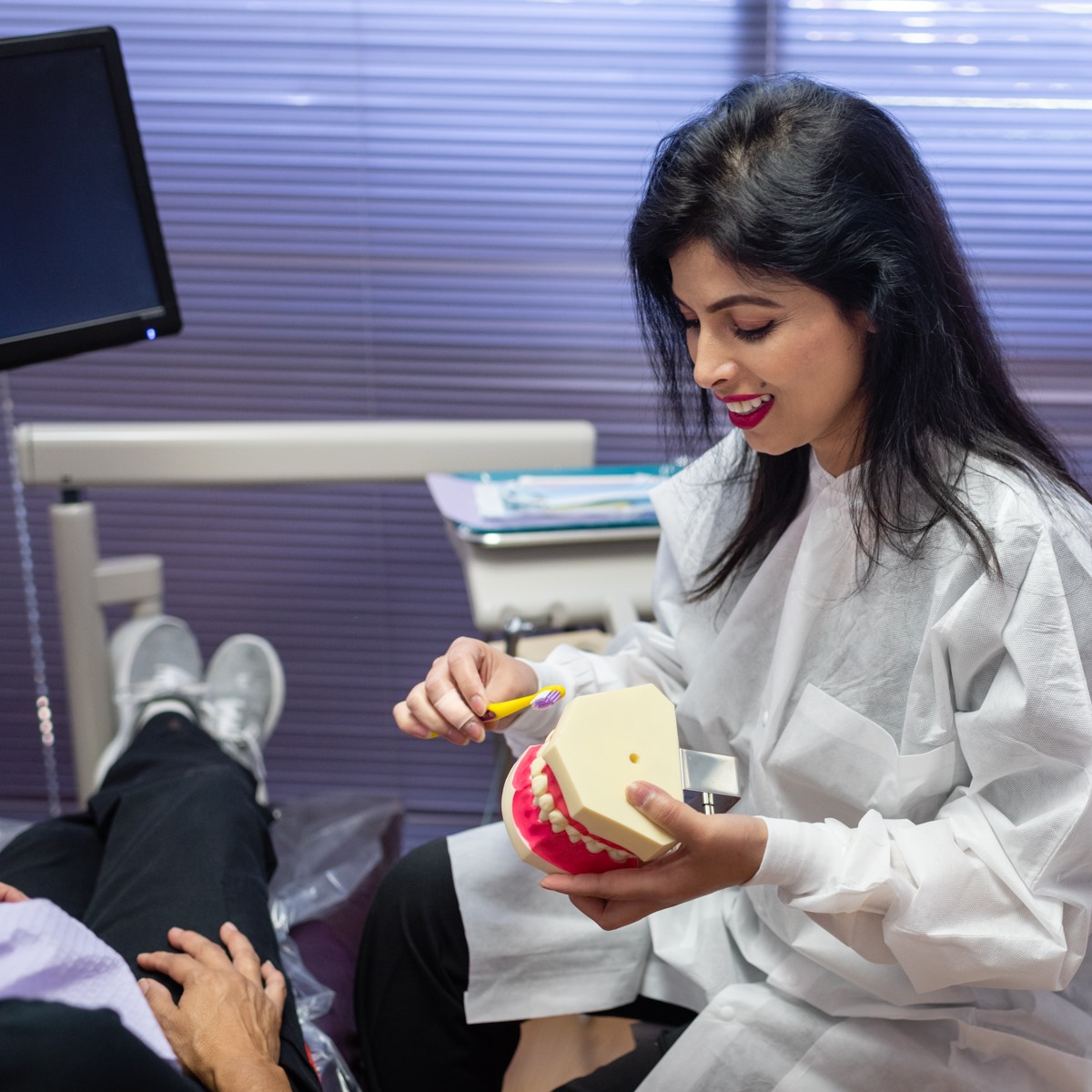 A cosmetic dentist in a white coat demonstrates tooth brushing technique on a dental model to a patient, highlighting the role of teeth whitening and oral care in achieving a bright, healthy smile.