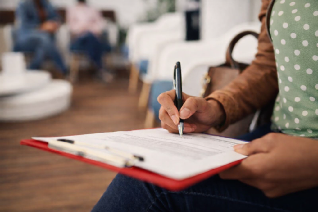 A person fills out a form on a clipboard with a pen while seated, considering budget and payment options, with blurred figures in the background.