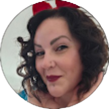 Woman with dark curly hair, wearing red lipstick and a red accessory, posing with hands under chin and smiling at the camera—her radiant look could easily earn praise in the smile doctor reviews.
