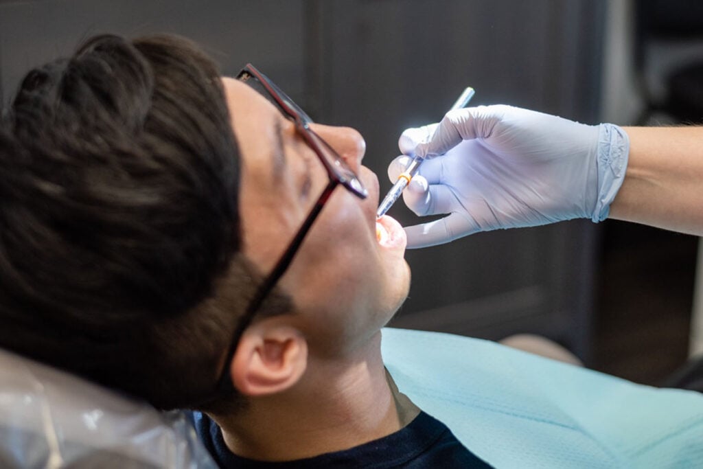 A person wearing glasses is reclining in a dental chair while a gloved dentist examines their mouth with dental instruments, performing subgingival curettage as part of advanced periodontics treatment in Sacramento.