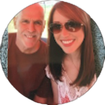 A man and a woman sit close together, smiling at the camera with warmth reminiscent of the smiles in the smile doctor reviews. The woman wears sunglasses and has long brown hair. The background is softly blurred.