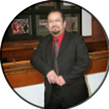 A man wearing a dark suit, red shirt, and patterned tie stands indoors in front of a wooden counter with framed pictures on the wall behind him, reminiscent of the friendly professionalism found in the smile doctor reviews.