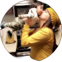 A woman in a yellow top and beanie lifts a small child and kisses their cheek in a kitchen setting, sharing a moment that could easily earn glowing the smile doctor reviews.