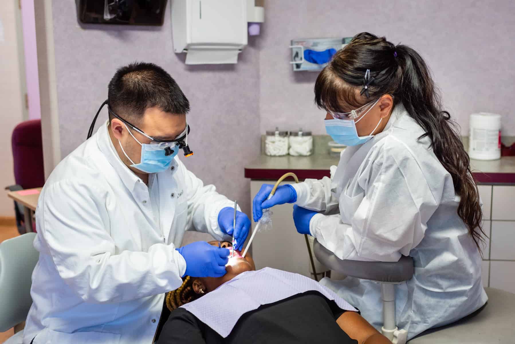 Two dental professionals wearing masks and gloves perform a dental procedure on a patient in need of an emergency dentist, lying in a dental chair.